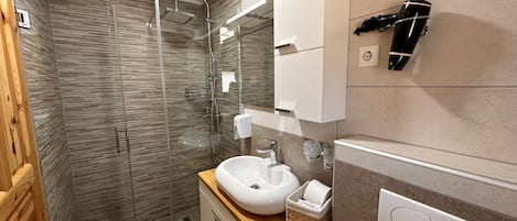A modern bathroom with a glass-enclosed shower, a wooden vanity, a white sink, and a hairdryer mounted on the wall.