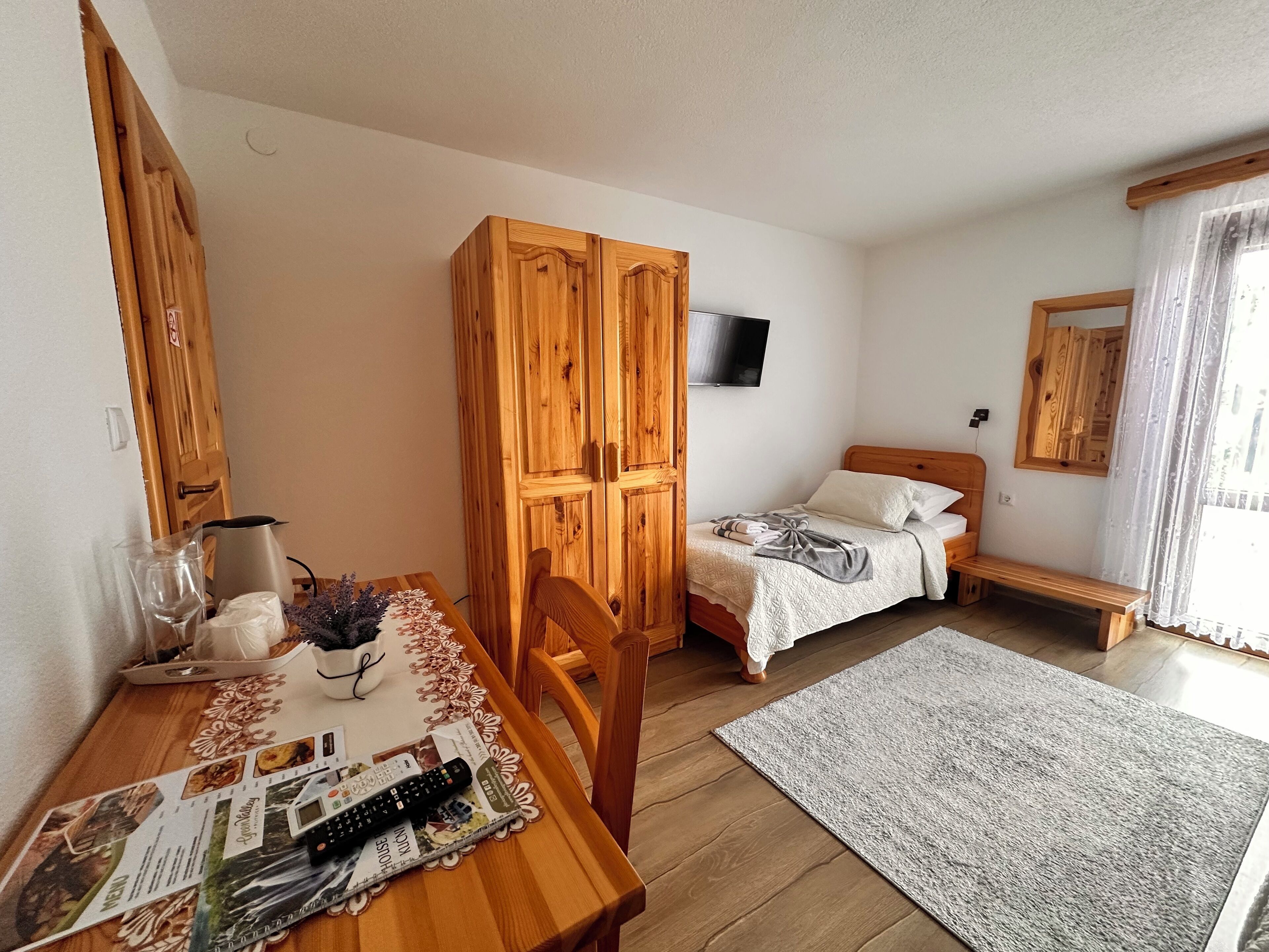 Una habitación con una mesa de comedor de madera, una cama con una manta gris, un armario de madera, un televisor montado en la pared y un banco junto a la ventana.