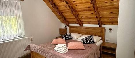 A bedroom with a sloped wooden ceiling, a wooden bed frame, a bedside table, and a window with curtains.