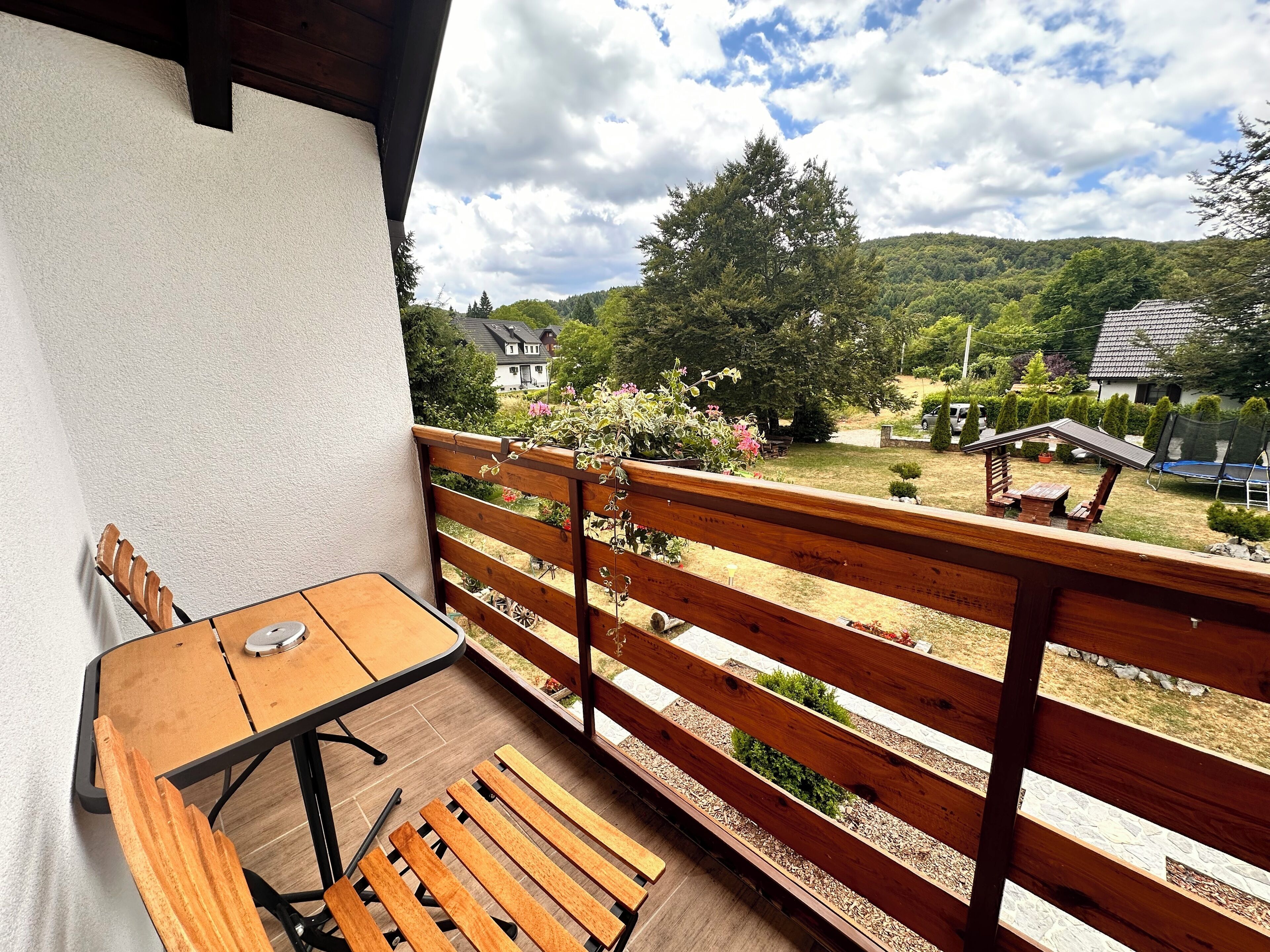 A balcony with a wooden bench and a table, overlooking a garden with a trampoline and a small shed.
