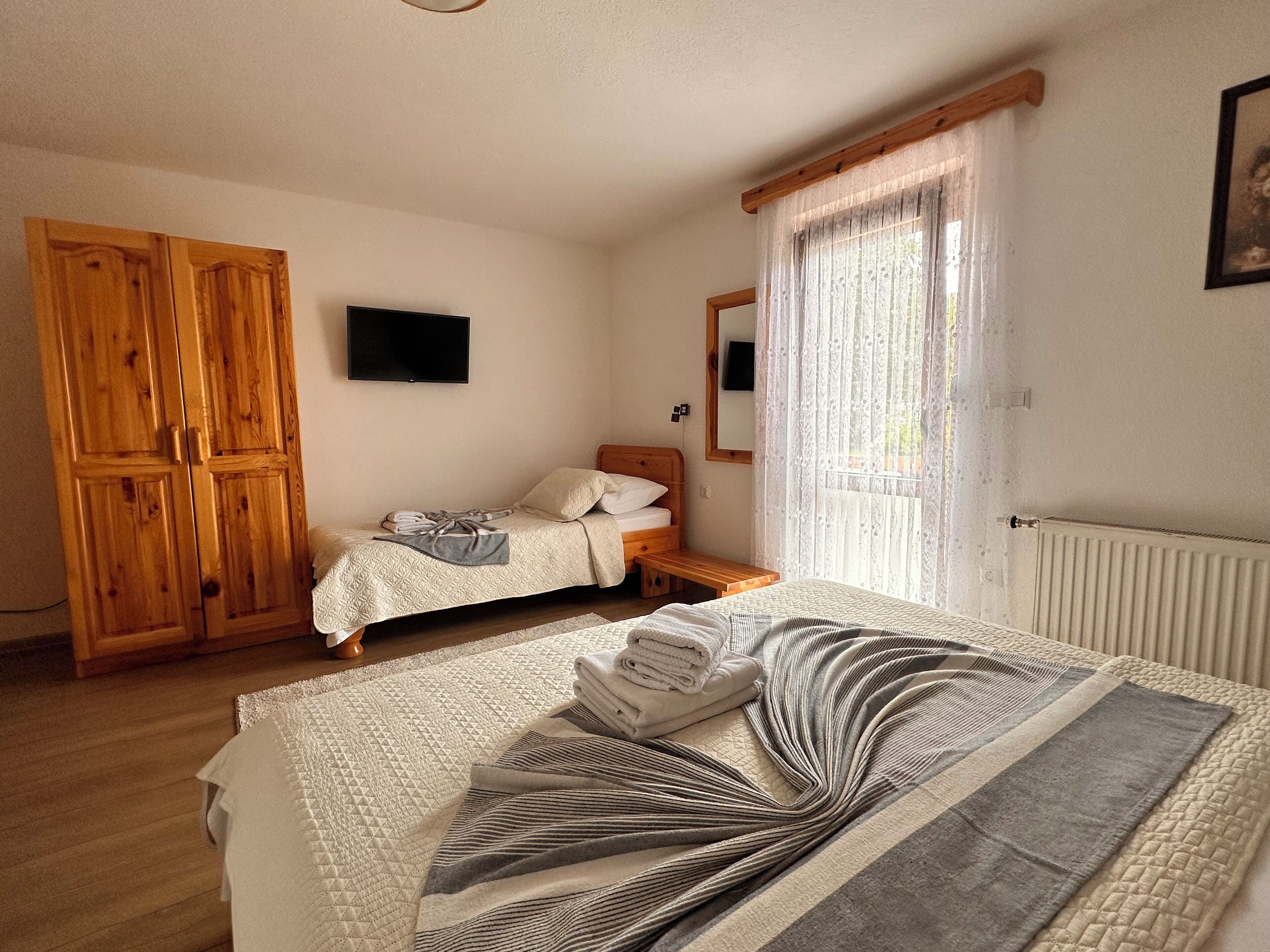 A bedroom with a large bed, a smaller bed, wooden wardrobe, a TV, and a window with curtains.