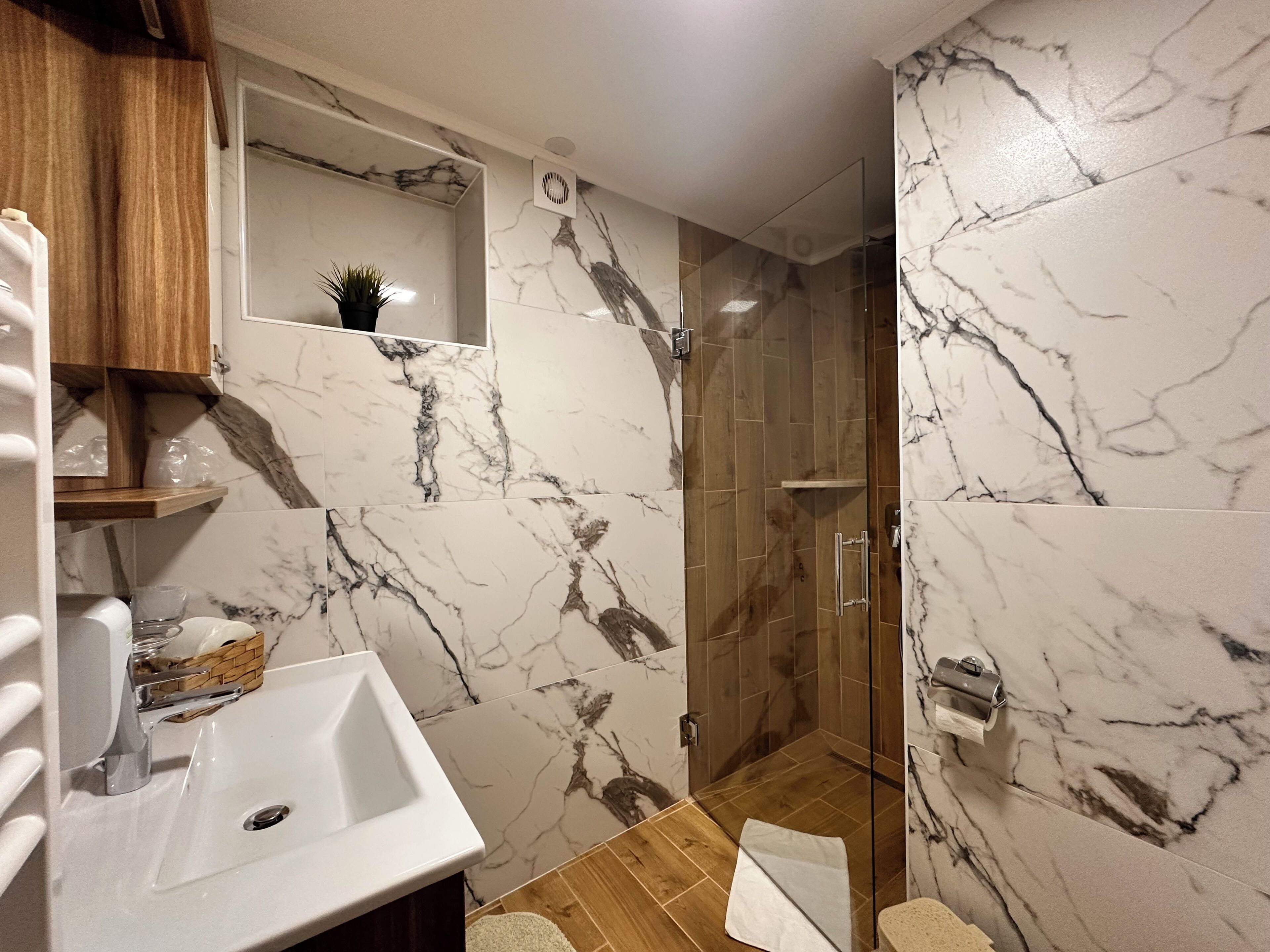 A bathroom with a white sink, wooden cabinets, a marble backsplash, and a glass shower enclosure.