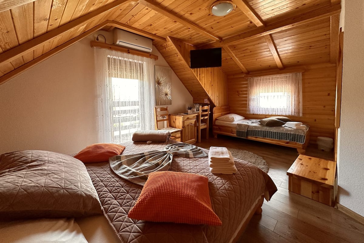 Un dormitorio acogedor en el ático, con techo inclinado, vigas de madera, una cama con colchas, un escritorio y una silla.