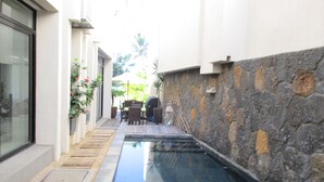 A narrow outdoor pool area with a stone wall on one side and a glass door on the other. There is a patio table and chairs, and a grill is visible in the background.