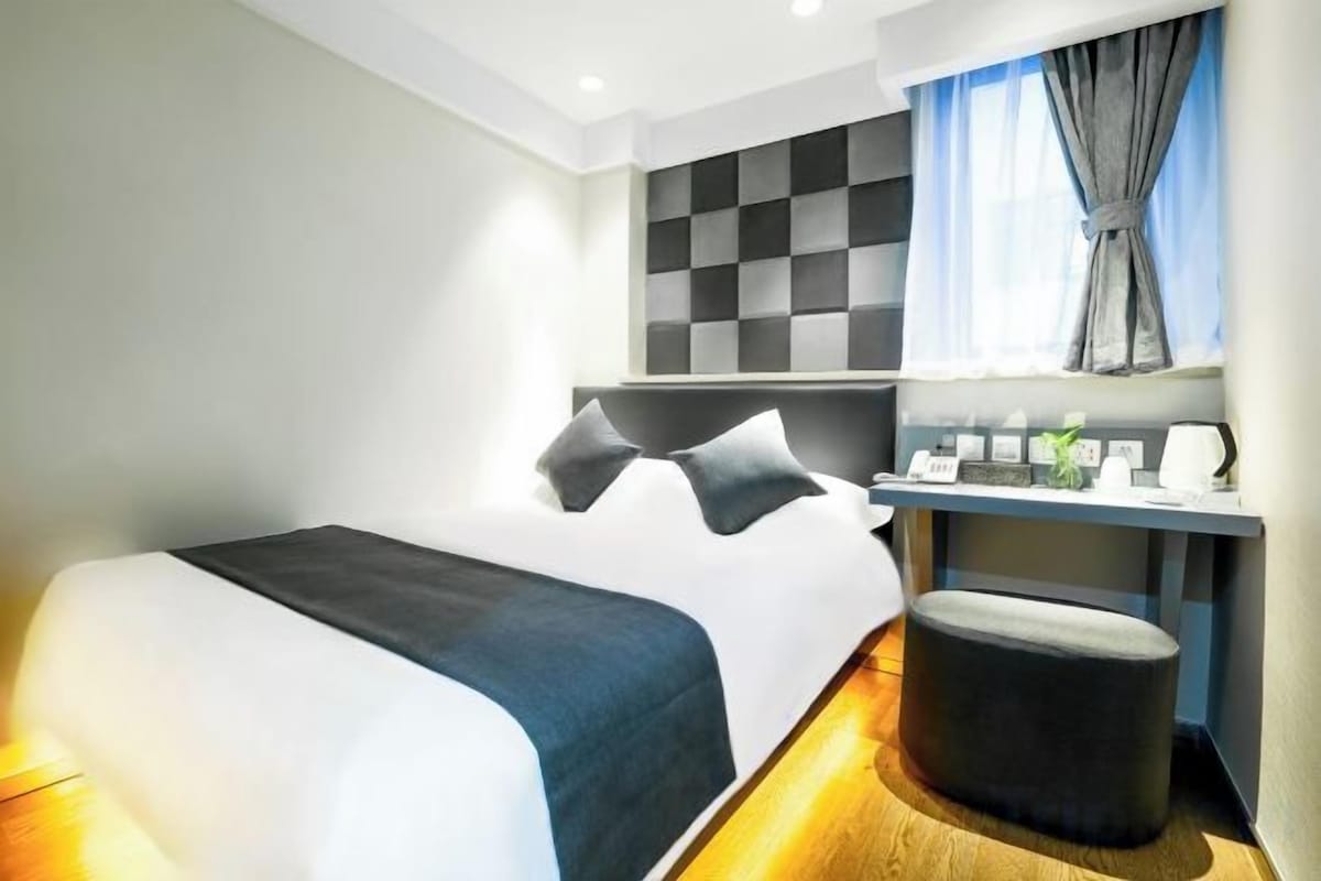 Habitación de hotel moderna con una cama grande, una mesita de noche, una pequeña zona de estar y una ventana con cortinas.