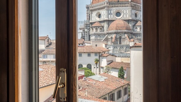 A view through a window of a building with a tiled roof and a dome structure in the background. - Florence Duomo View (Florence)