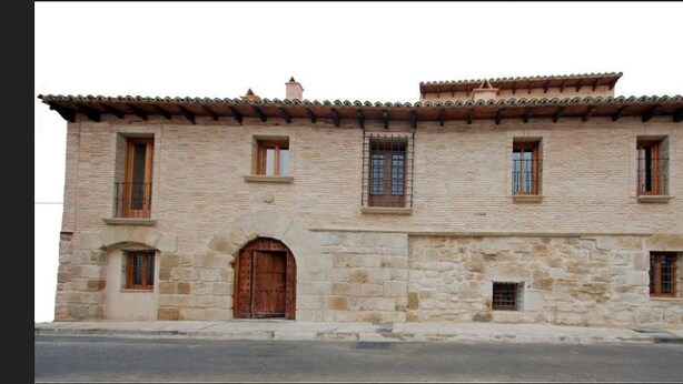 A two-story stone building with a central entrance, multiple windows, and a tiled roof. - A Corona del Reino (Ejea De Los Caballeros)