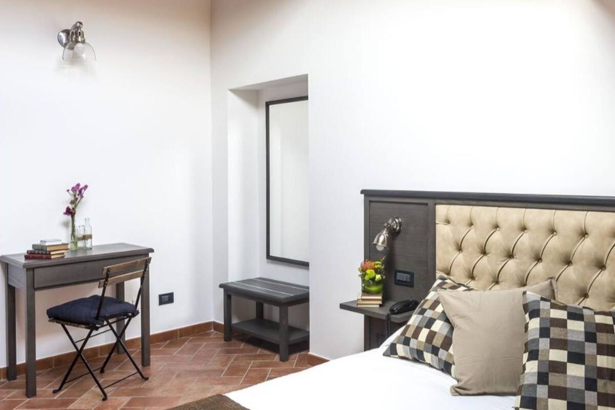 A bedroom with a bed, a desk, a chair, a small table, a mirror, and a nightstand.