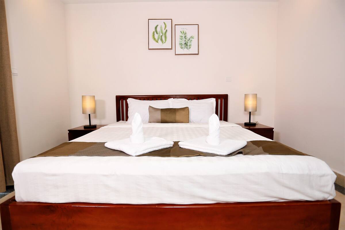 A neatly made bed with white linens and two towels folded at the foot. There are two bedside tables with lamps, and two framed botanical prints on the wall.
