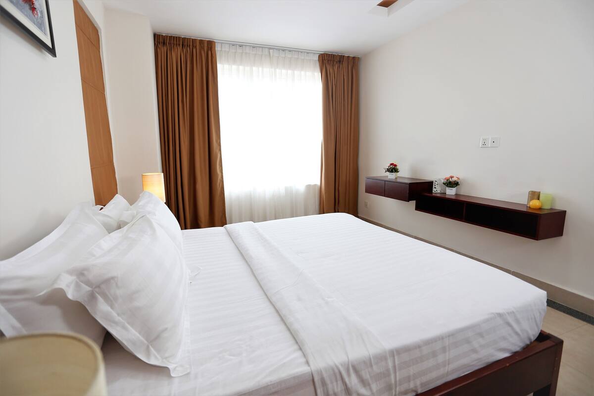 A neatly made bed with white linens, a bedside lamp, and a shelf with decorative items.