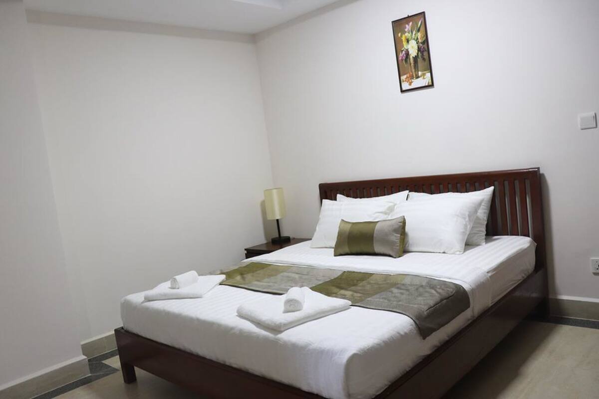A neatly made bed with white linens and a brown headboard, a small bedside table with a lamp, and a framed picture on the wall.