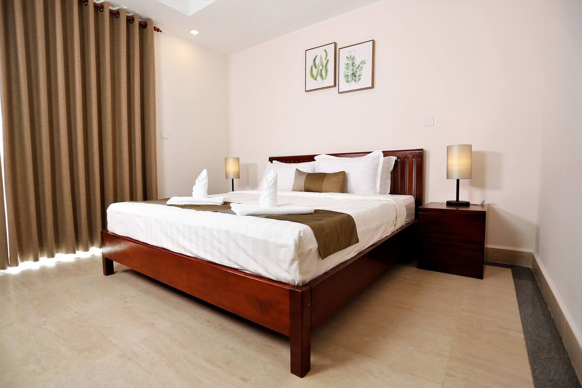 A neatly made bed with white linens and a brown throw, a wooden headboard, two bedside lamps, and two framed botanical prints on the wall.