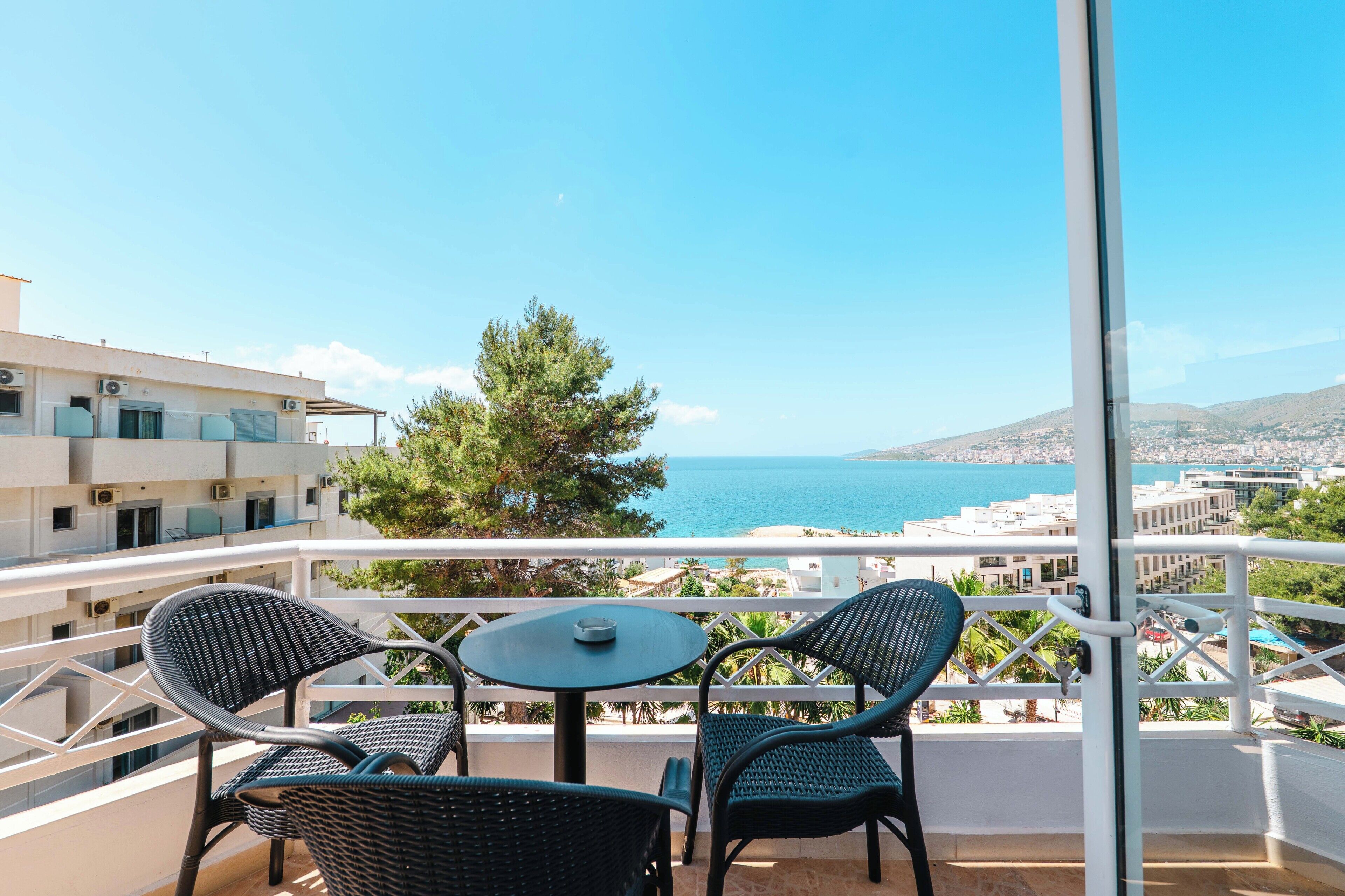 A balcony with two wicker chairs and a small round table, overlooking a coastal cityscape with clear skies.