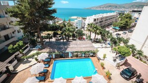 Zona de piscina amb gandules i ombrelles, vista al mar i edificis circumdants.