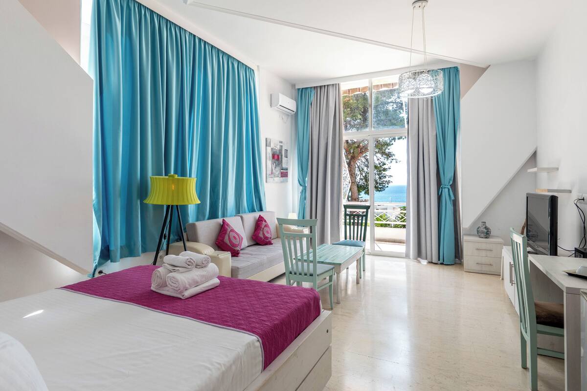 Habitación de hotel con cama, sofá, escritorio y silla. Cuenta con ventana con cortinas, lámpara y vistas al exterior.