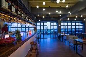 Zona de bar con vistas a la ciudad a través de amplios ventanales, suelo de madera y lámparas colgantes.