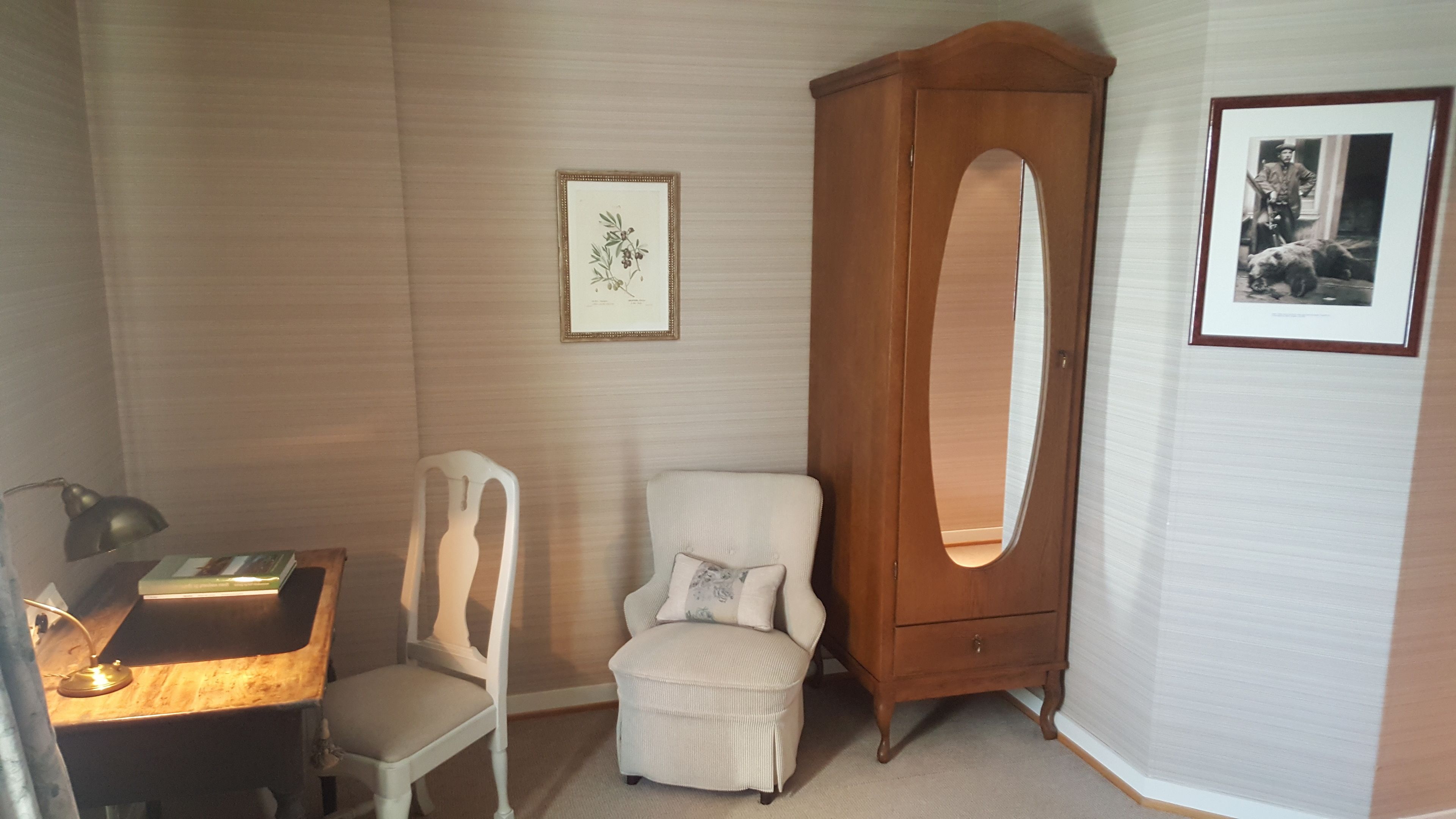 Um quarto pequeno com um armário de madeira, uma cadeira branca, uma mesa com uma luminária e duas fotos emolduradas na parede.