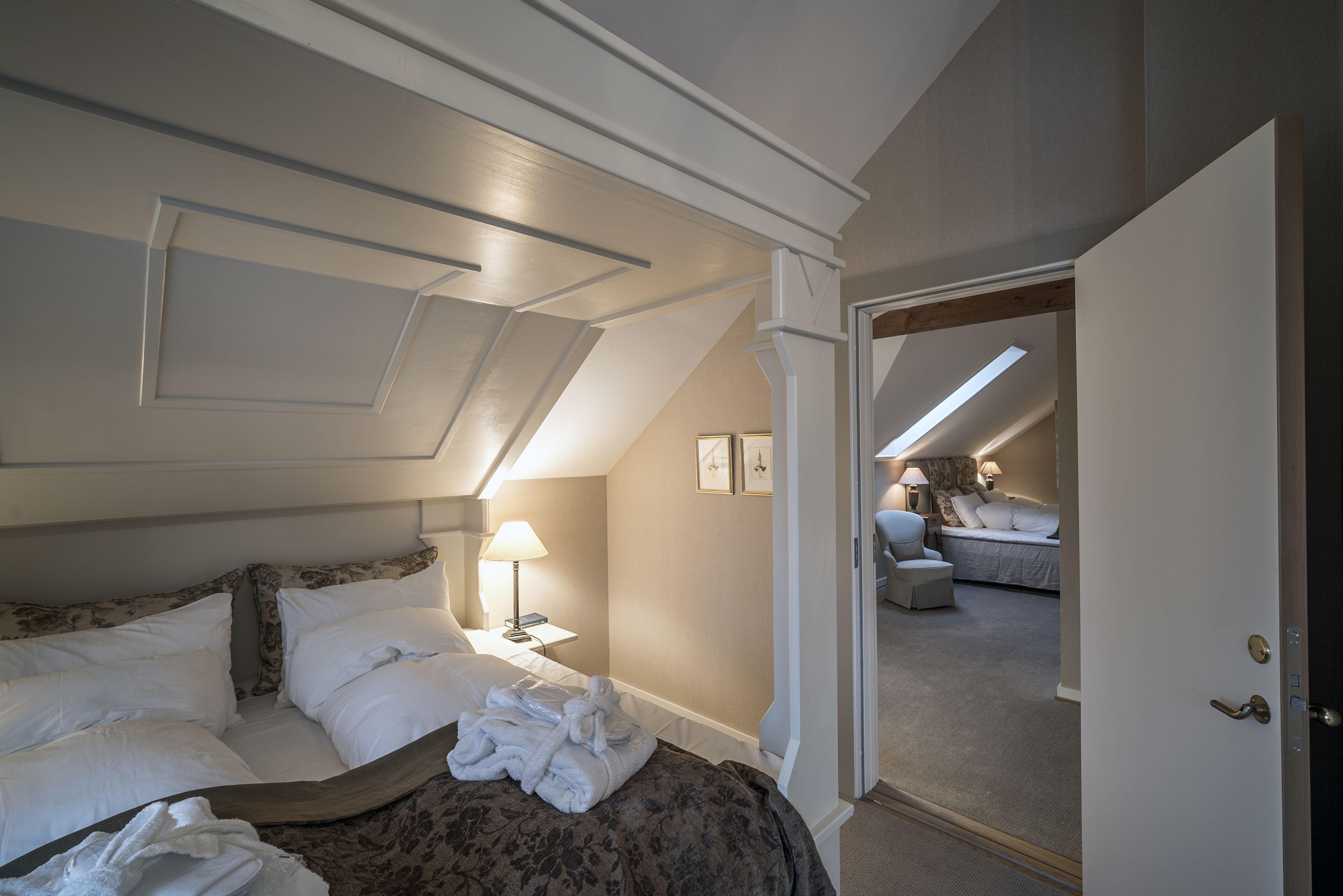 Een hotelkamer met een bed, een nachtkastje met een lamp, en uitzicht op een andere kamer door een geopende deur.
