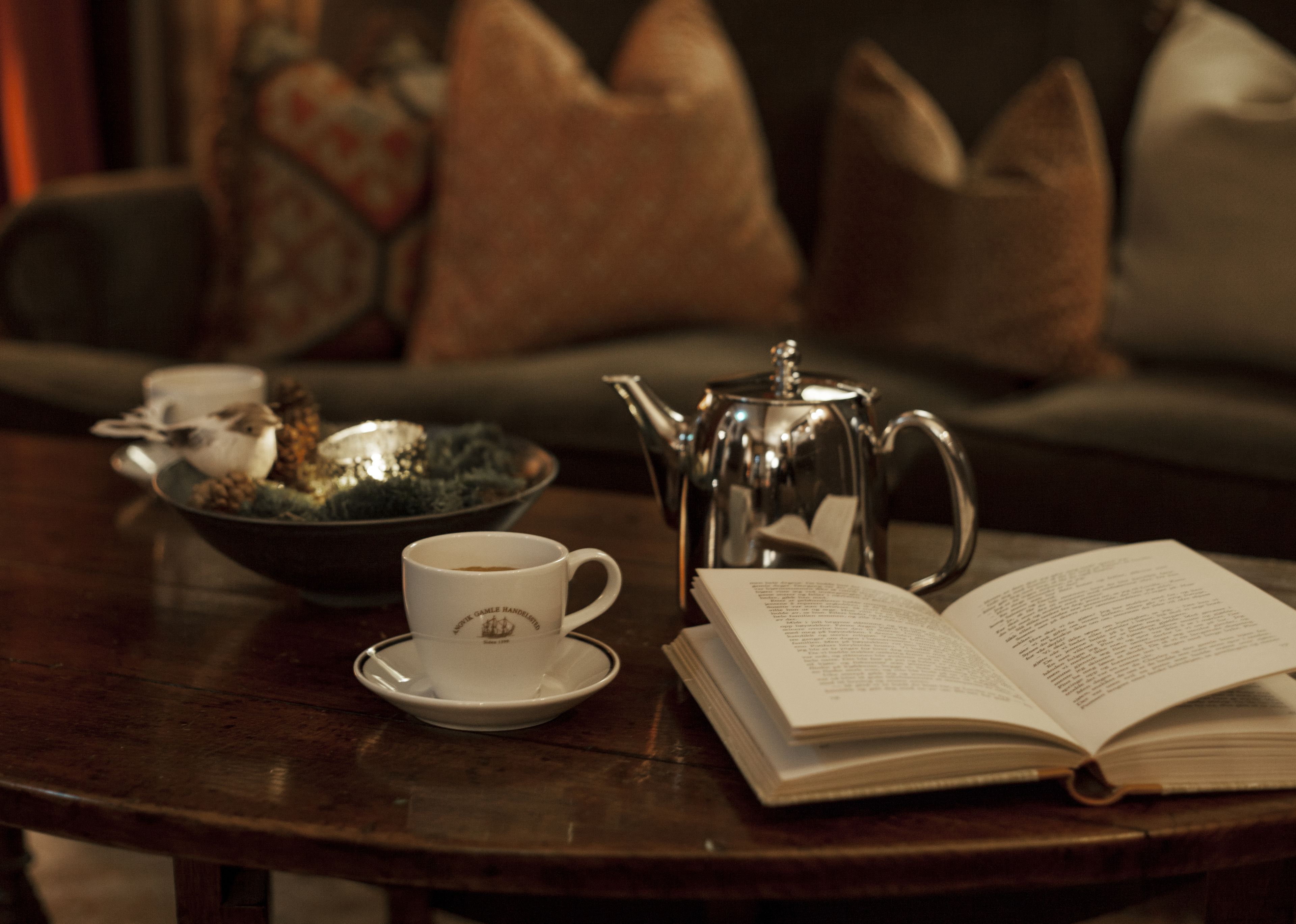 Una mesa de madera con una taza blanca, una tetera plateada y un libro abierto.