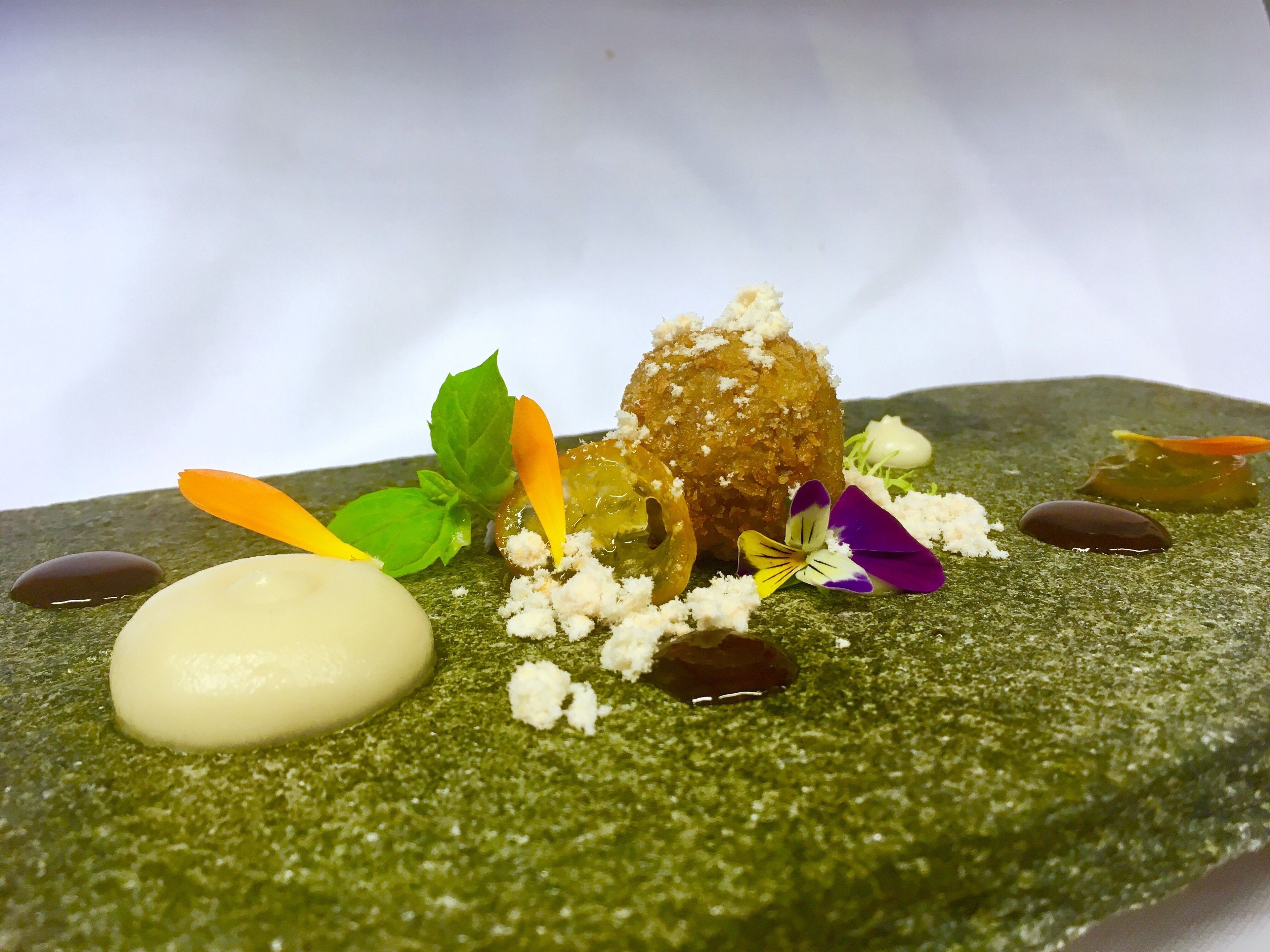 Un plato de postre con una base verde texturizada, un elemento blanco esférico y varios adornos, incluyendo una hoja, una flor y una rodaja pequeña de naranja.
