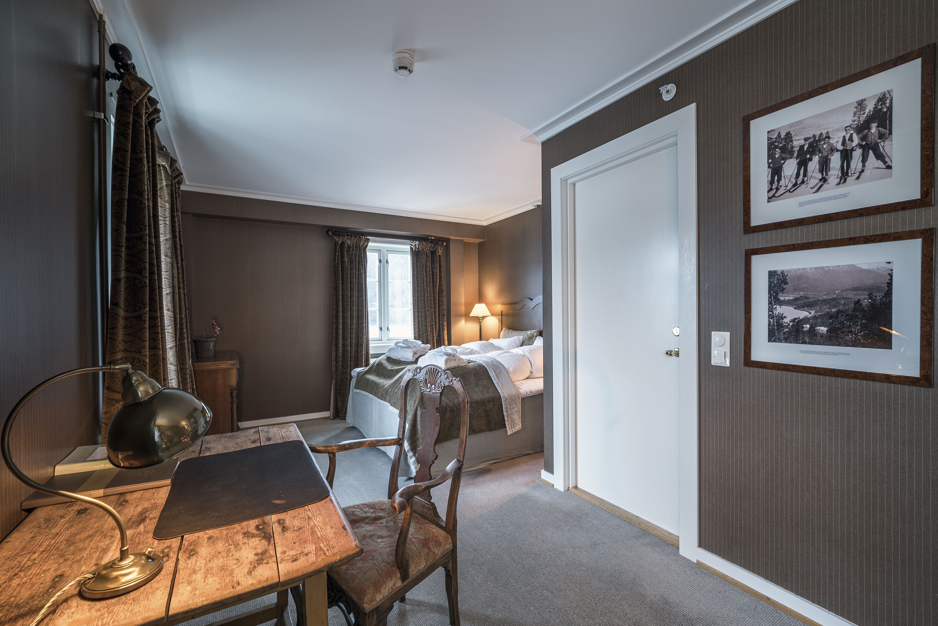 Una camera d'albergo con una scrivania in legno, una sedia, un letto con un comodino, una lampada e due quadri appesi al muro.