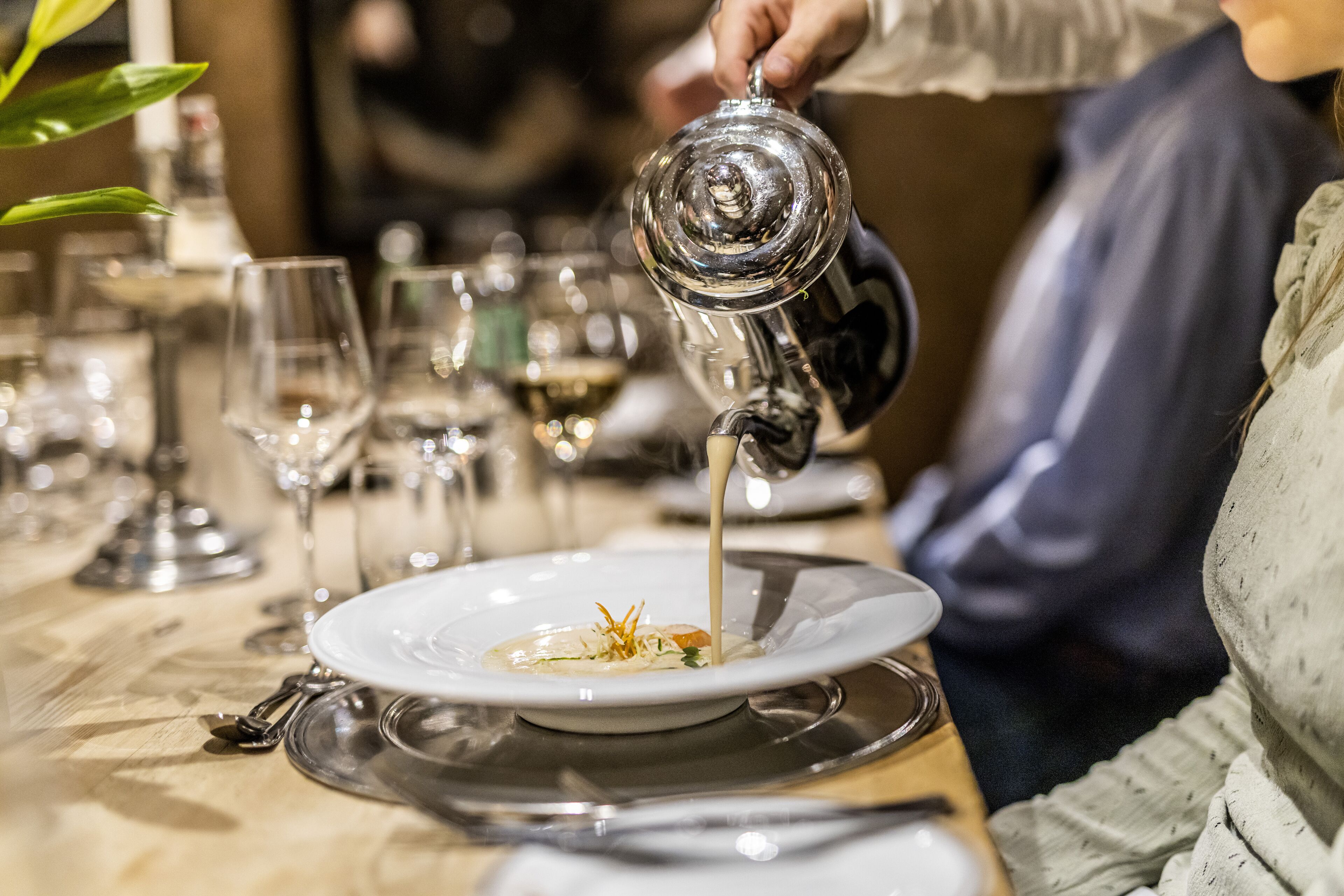 A person pouring a clear liquid from a metal pitcher onto a white plate with a garnish, with glasses and cutlery on the table.