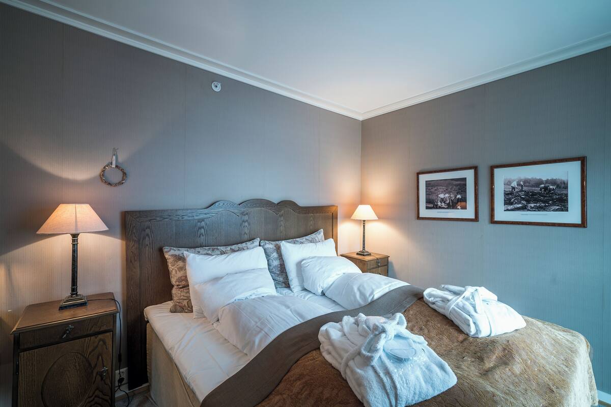A neatly made bed with white linens, a wooden headboard, and two bedside lamps. There are two framed pictures on the wall and a small wooden nightstand with a lamp.