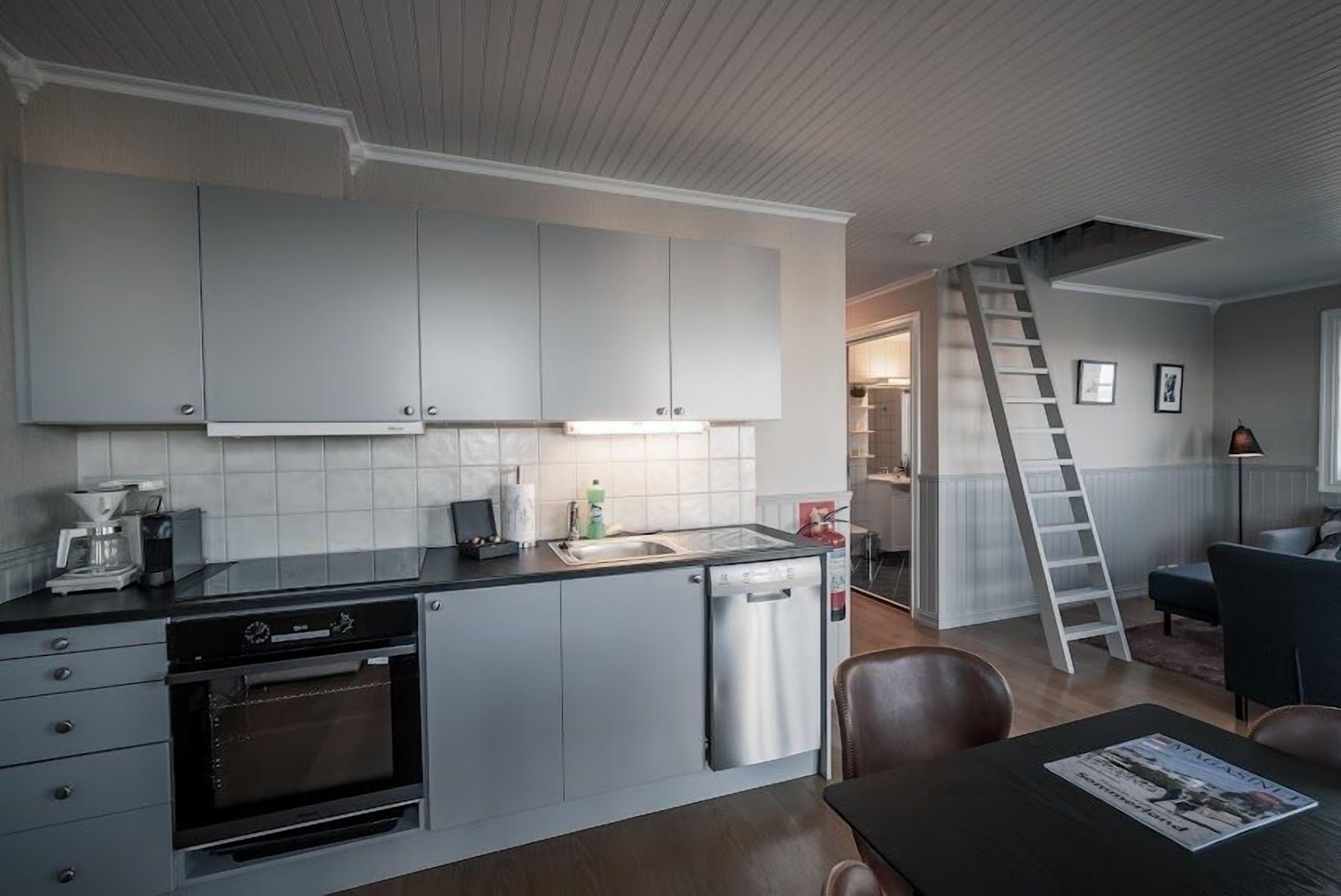 A modern kitchen with grey cabinets, a dishwasher, and a built-in oven. There is a dining table with chairs and a ladder leading to a lofted area.