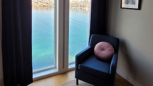 A room with a dark blue armchair, a round woven rug, and a view of a rocky shoreline through large glass doors.