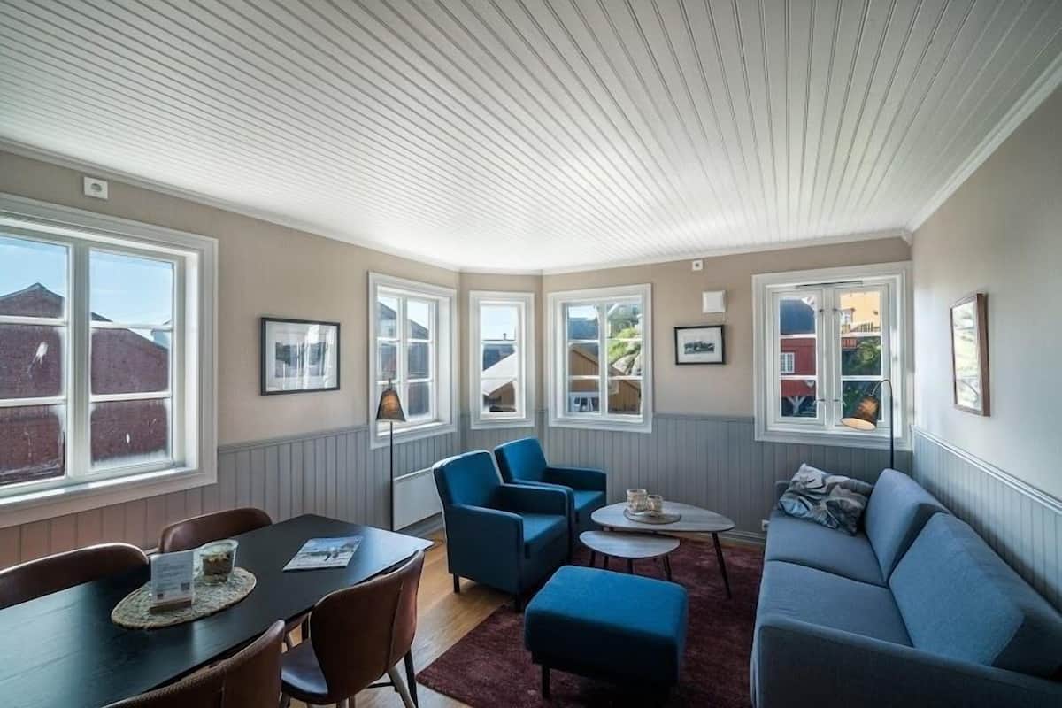 A cozy living room with a dark wooden table, blue armchairs, a grey sofa, and a small coffee table. Large windows provide natural light and views of the outside.