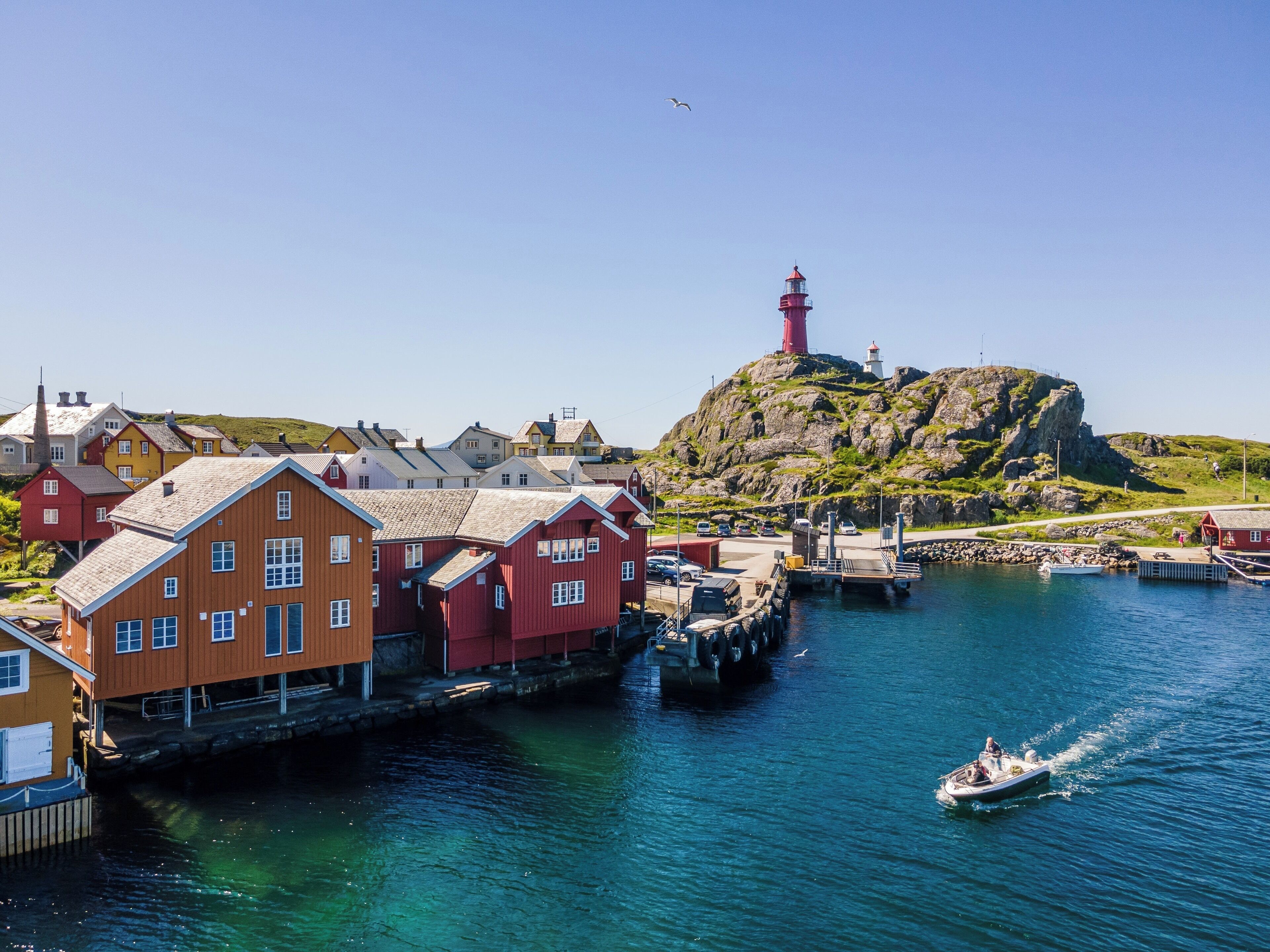 A coastal town with colorful wooden houses, a lighthouse on a rocky hill, and a boat on the water.