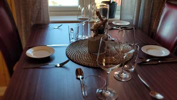 A dining table set with plates, cutlery, and wine glasses, with a potted plant and a window in the background.