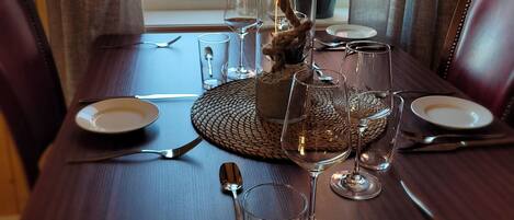 A dining table set with plates, cutlery, and wine glasses, with a potted plant and a window in the background.