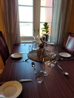 Ein Esstisch ist gedeckt mit Tellern, Besteck und Weingläsern. Im Hintergrund steht ein Blumentopf und ein Fenster ist zu sehen.