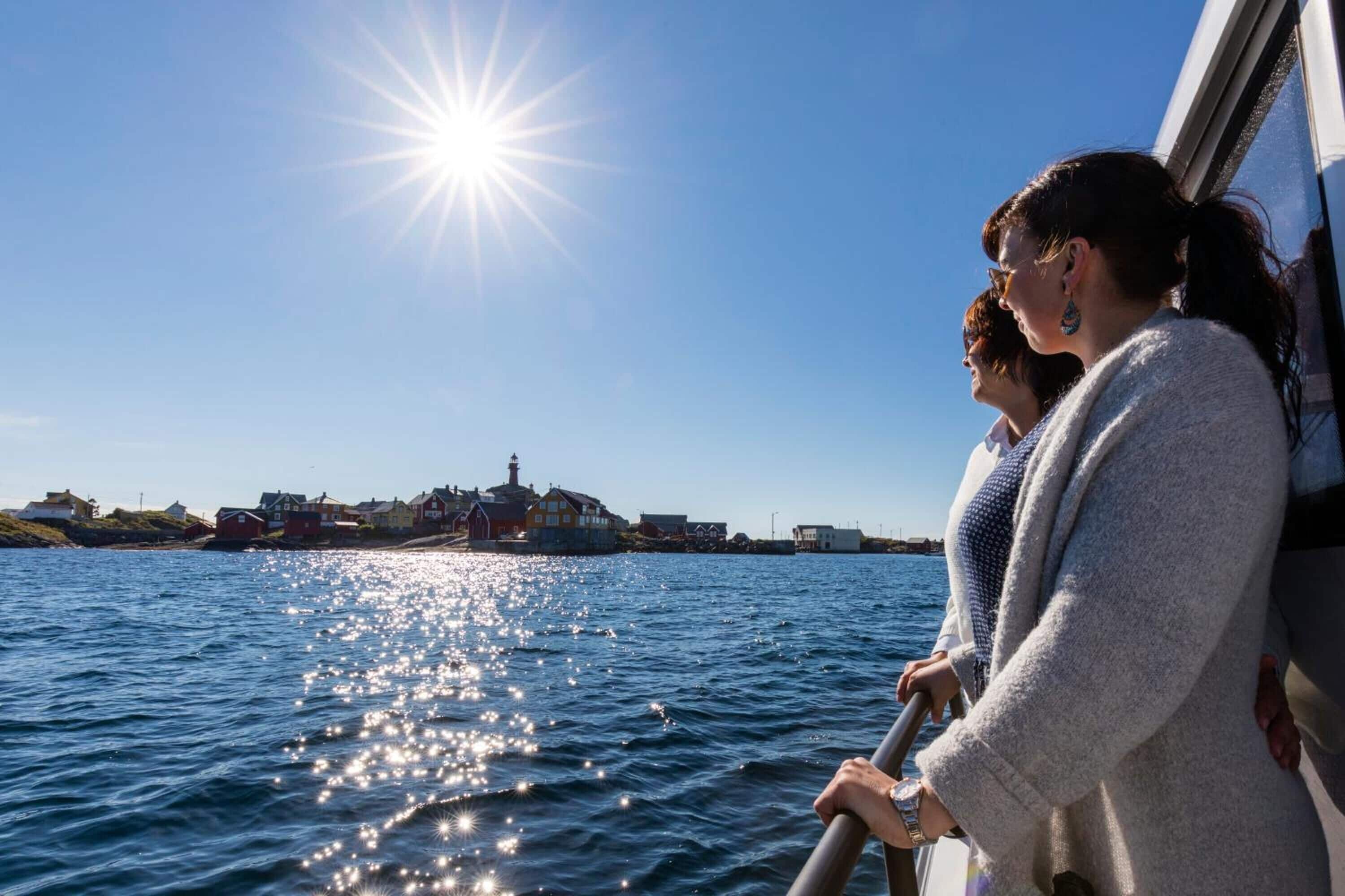 A boat with two passengers looking at a coastal town with buildings and a lighthouse.