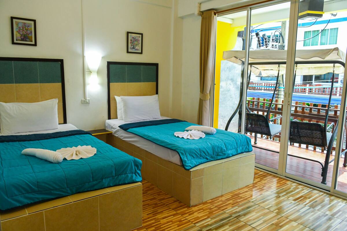 Two beds with teal bedding and white pillows, a wooden floor, and a view of a balcony with outdoor furniture.