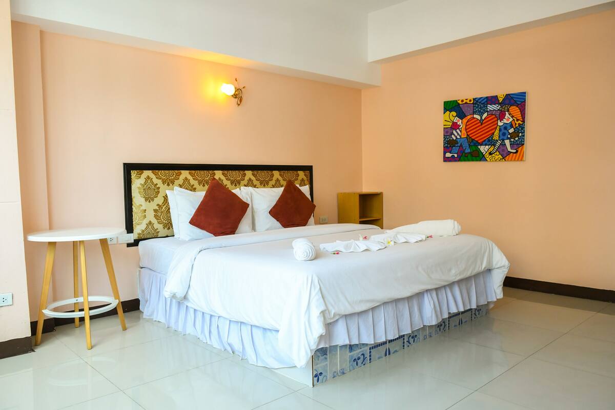 A neatly made bed with white linens and brown pillows, a small round table, and a colorful painting on the wall.