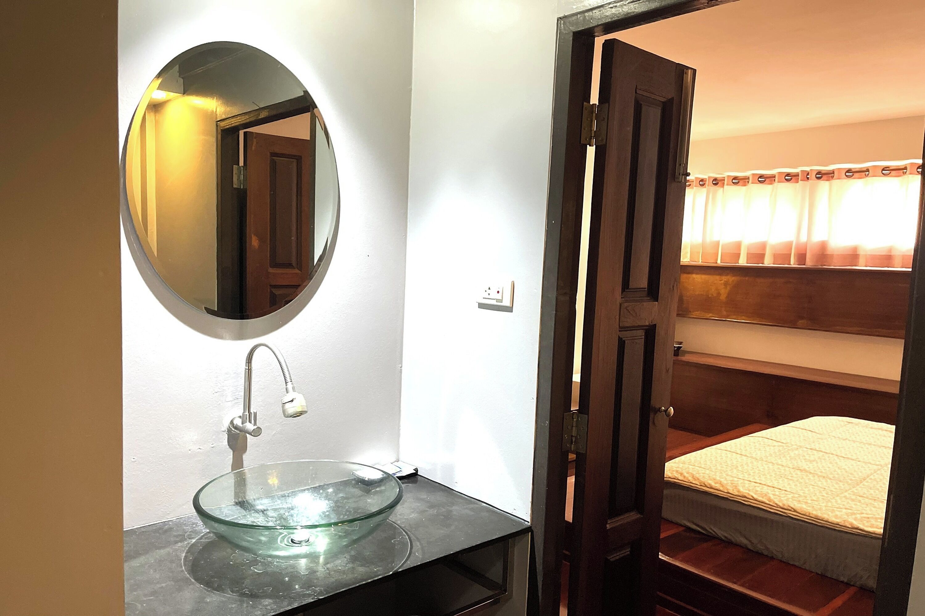 Un baño con un lavamanos de vidrio, un espejo redondo y una puerta de madera que da a una habitación con literas.