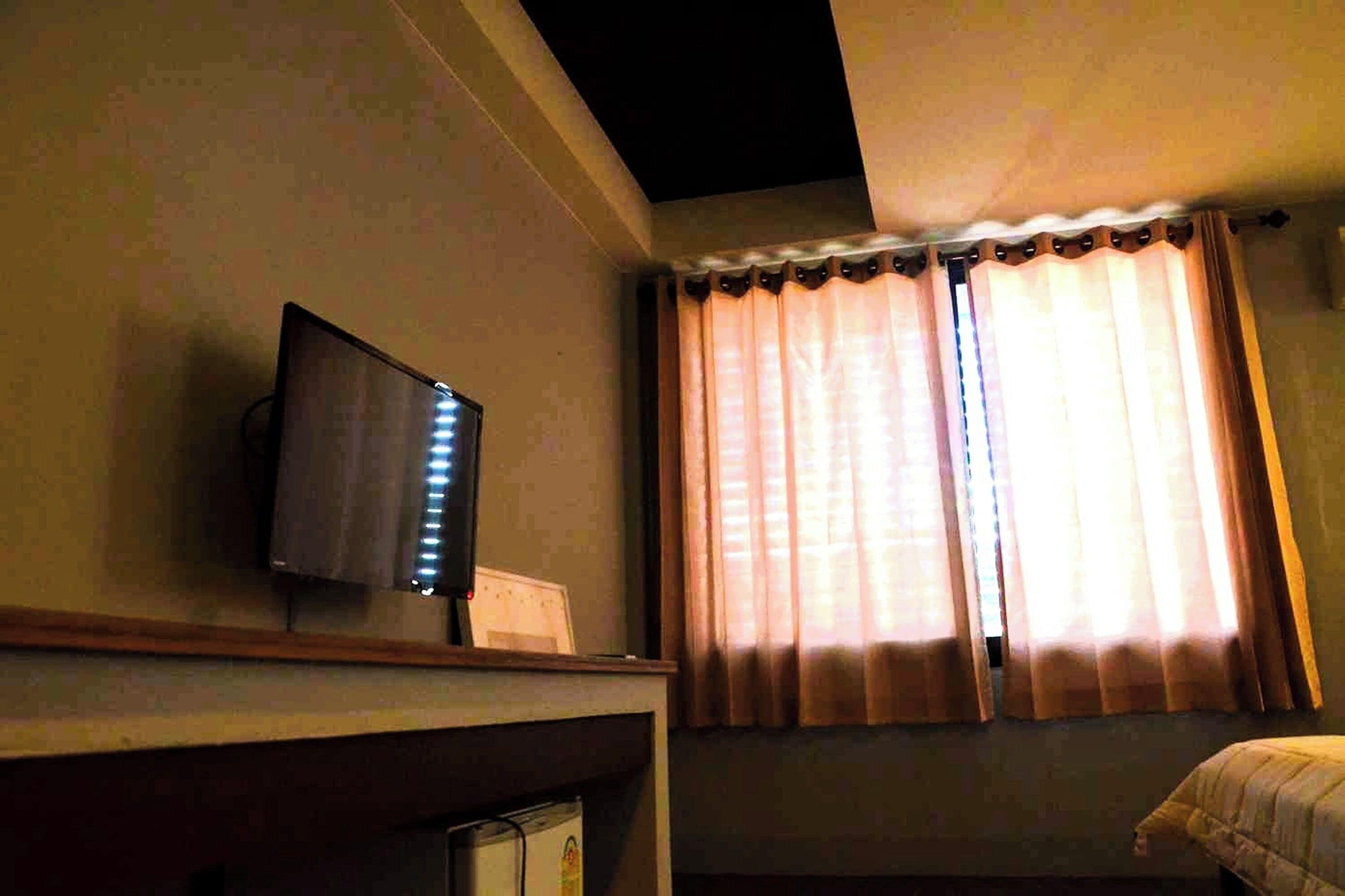 A room with a television on a stand, a window with curtains, and a bed.