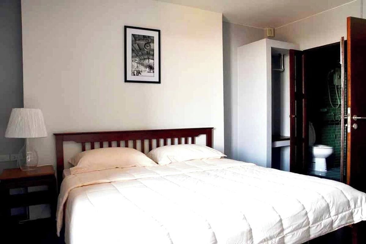 A neatly made bed in a hotel room with a framed picture on the wall, a bedside lamp, and an open door leading to a bathroom.