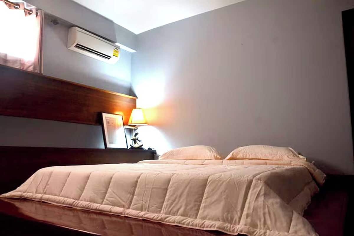 A bedroom with a bed, an air conditioner, a nightstand with a lamp, and a wall-mounted shelf.