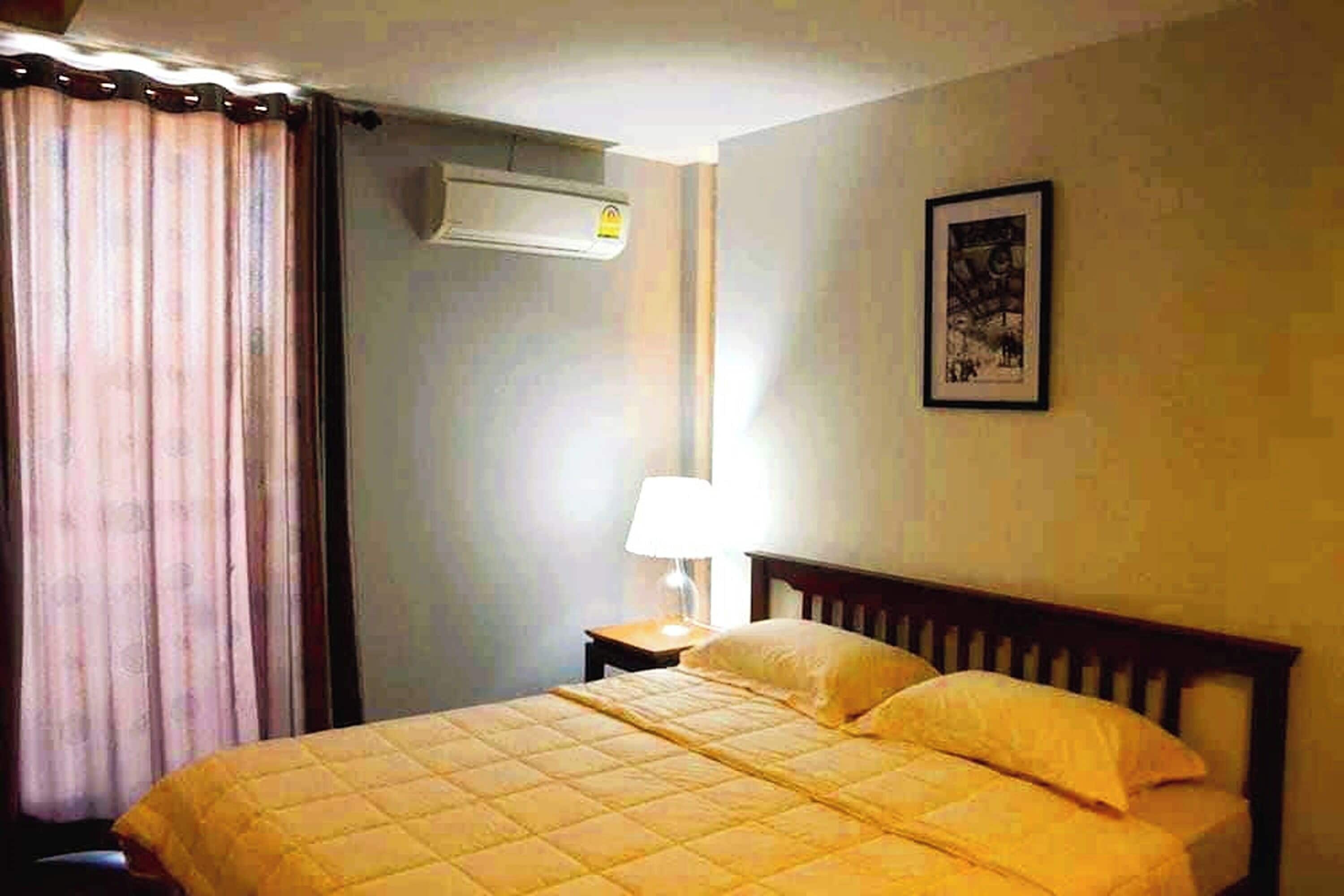 A hotel room with a bed, a nightstand, a lamp, a picture on the wall, and an air conditioning unit.