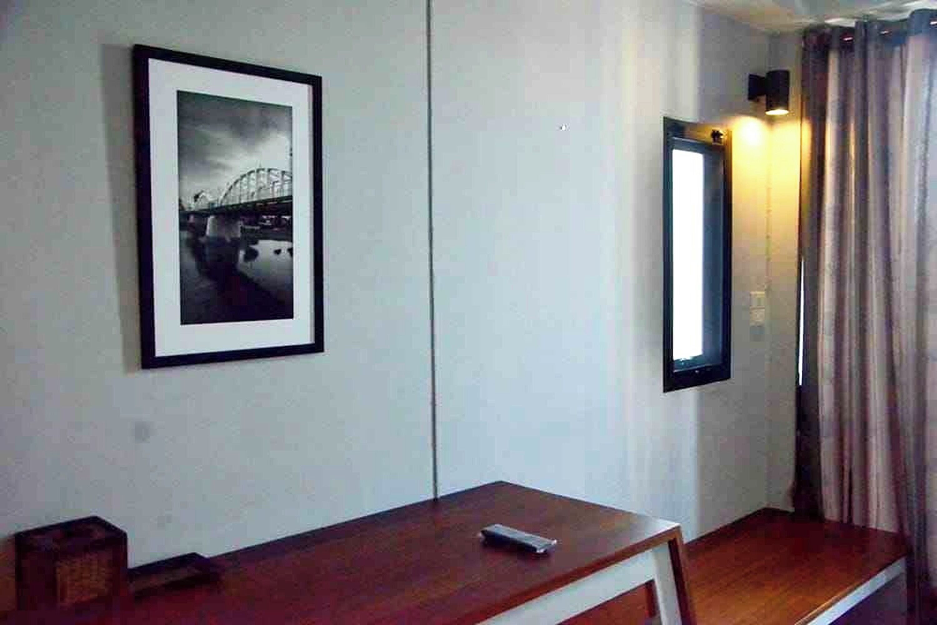 A hotel room with a framed picture of a bridge, a bedside table with a remote control, and a mirror reflecting a curtain.