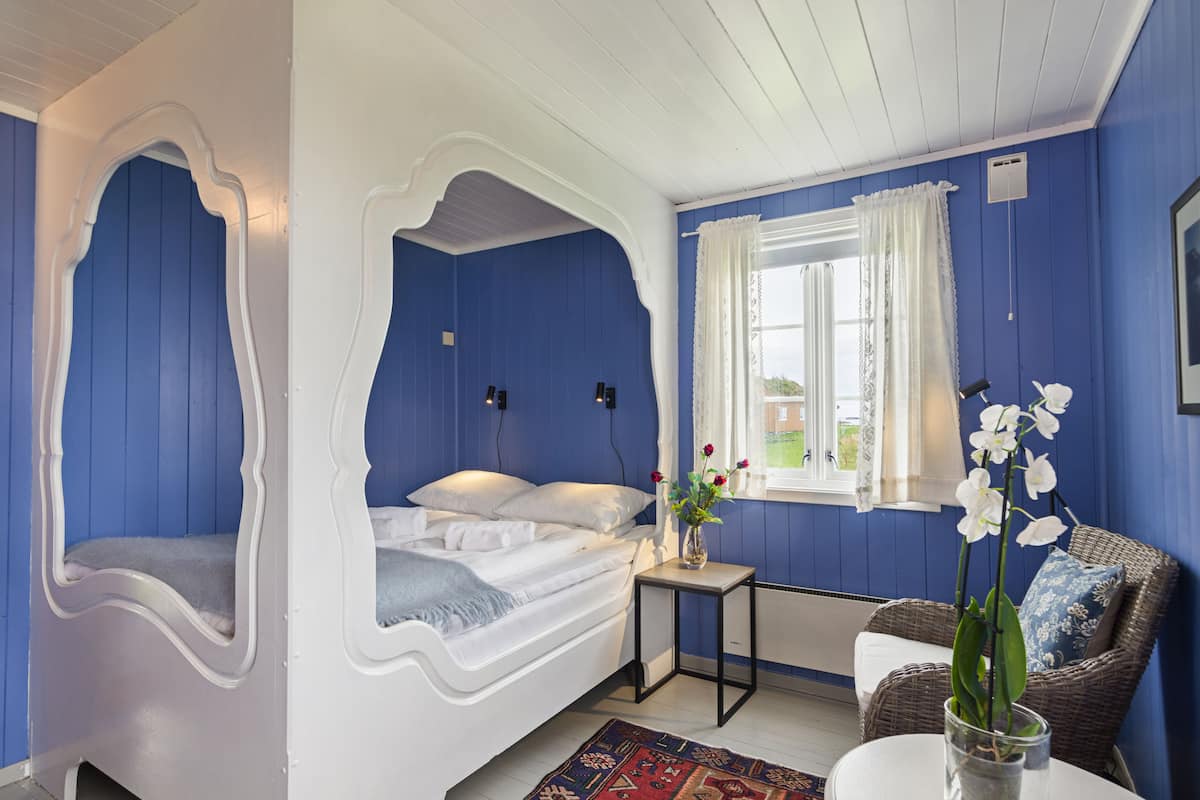 Un dormitorio con una cama blanca, una mesita con un florero, una silla de mimbre y una ventana con cortinas.