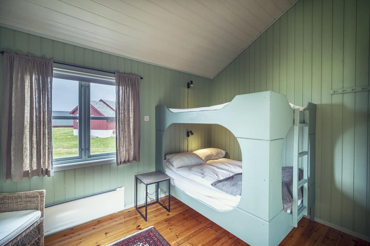 Una cama litera con escalera, una mesita pequeña y una ventana con cortinas.