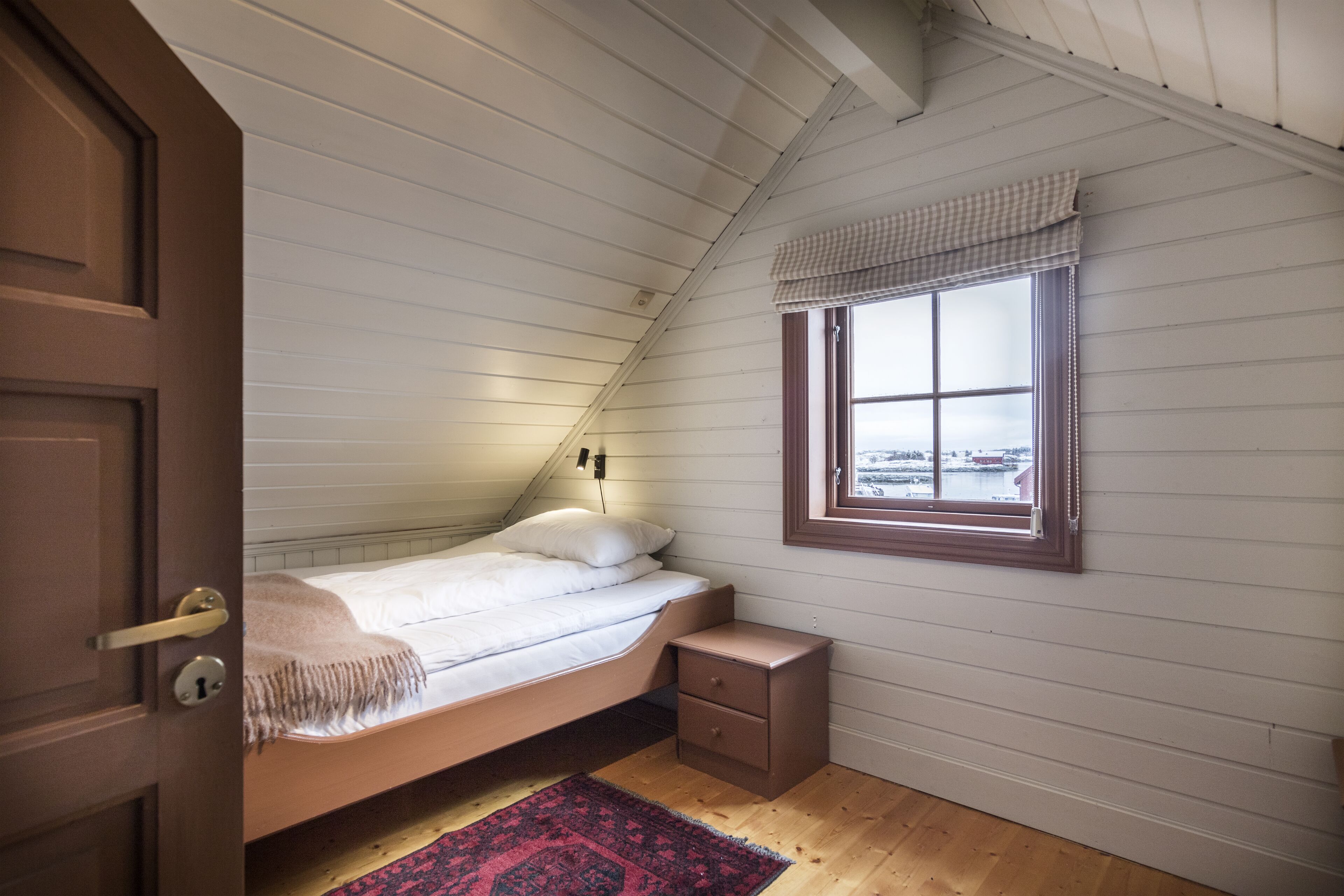A small, single-bed room with a wooden floor, a window with a checkered valance, and a nightstand.