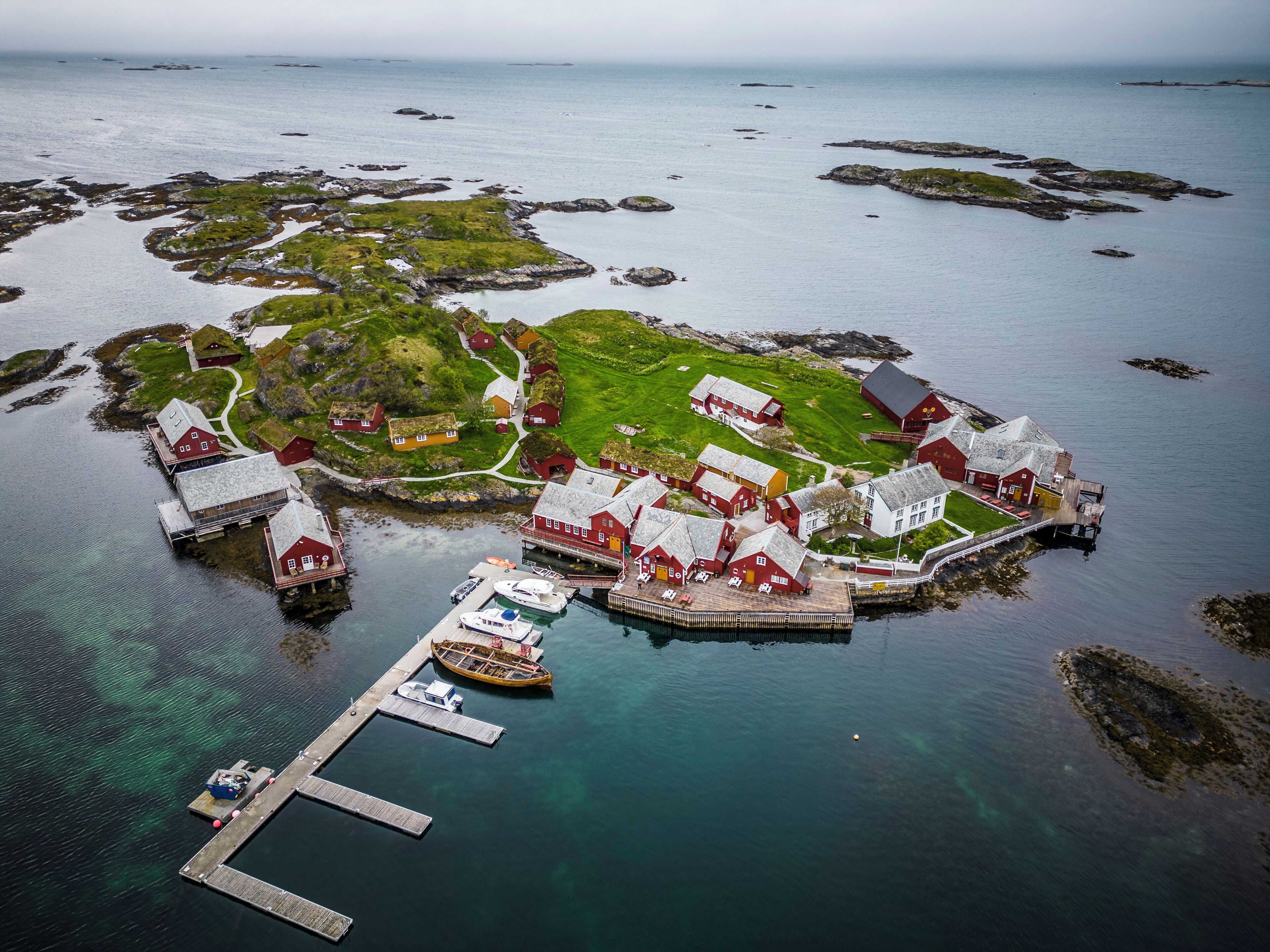 An aerial view of a coastal village with traditional red-roofed buildings, a dock with boats, and clear blue waters.