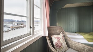 A room with a wicker chair, a bed with a green blanket, and a view of a dock with a boat. - Håholmen Havstuer - by Classic Norway Hotels (Averoy)