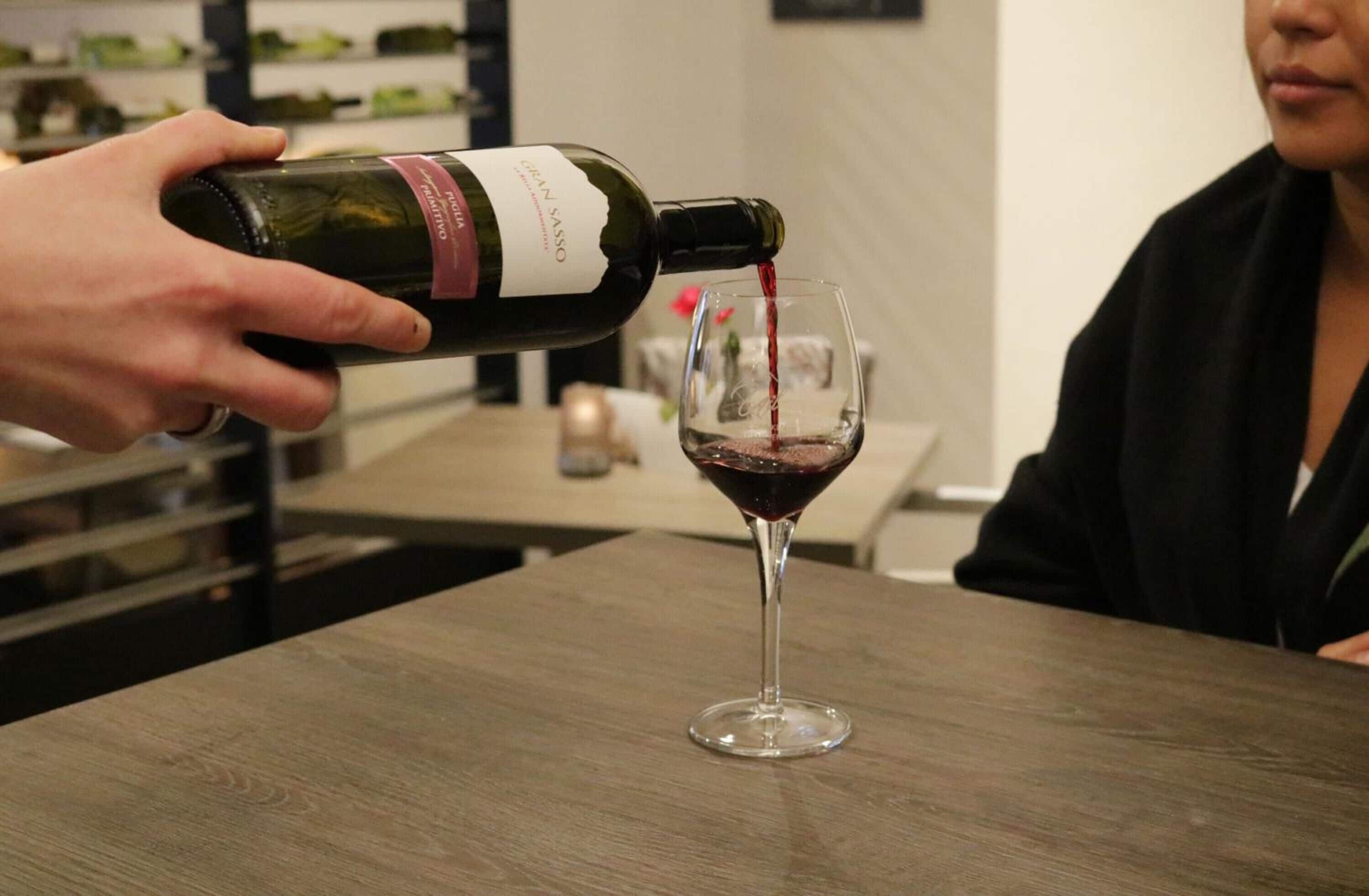 A person pouring red wine from a bottle into a glass on a wooden table.