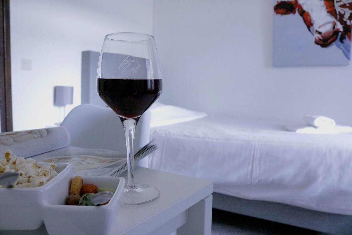 A glass of red wine on a white table with a bed and a painting in the background.
